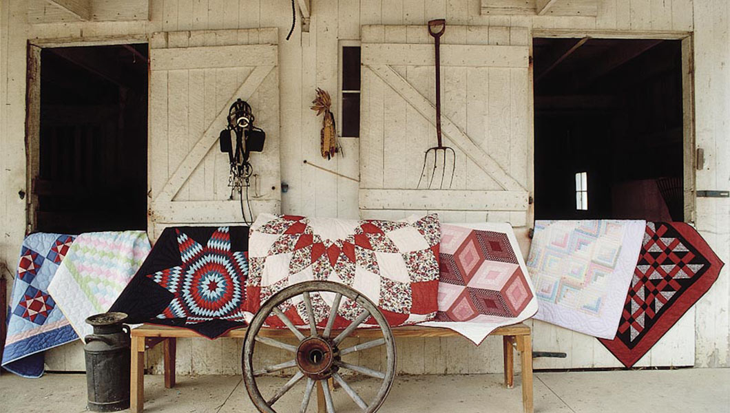 Amish Quilts in front of a barn with wagon wheel